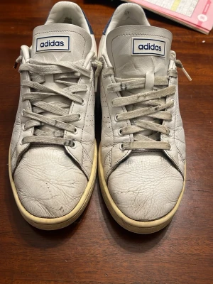 Adidas Stan Smith vita sneakers - Klassiska Adidas Stan Smith sneakers i vitt skinn med blå detalj på hälen. Skorna har perforerade sidor, rund tå och snörning framtill. Sulan är beige och har ett hexagonmönster undertill. Perfekta för dig som gillar retrostil och tidlös design.