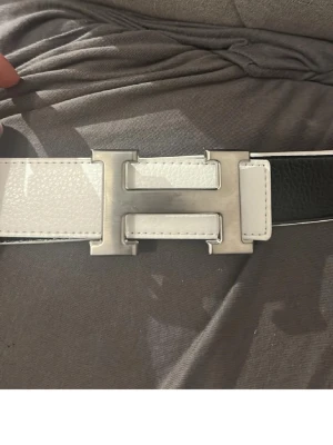 Svart och vit skärp med H-spänne - Säljer ett stilrent skärp i svart och vitt med ett stort metallspänne i form av ett H. Skärpet har en klassisk design med kontrasterande färger och vita sömmar längs kanterna. Perfekt accessoar för att lyfta vilken outfit som helst.