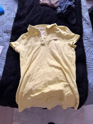 Gul piké från Hollister - Säljer en ljusgul pikétröja från Hollister med klassisk krage och knappslå framtill. Tröjan har korta ärmar och den ikoniska Hollister-loggan broderad på bröstet. Tillverkad i mjuk bomull, perfekt för en avslappnad och fräsch look.