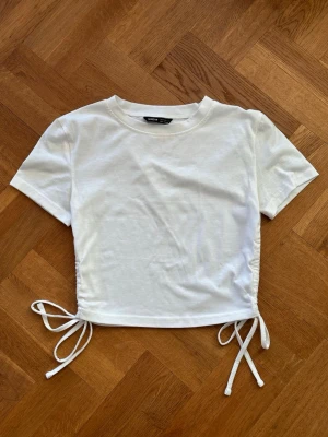 Vit croppad topp med knyt  - Säljer en vit croppad topp från SHEIN med korta ärmar och rund hals. Toppen har knytband i sidorna och gör att du kan justera passformen. Perfekt till jeans eller kjol för en enkel men stilren outfit.