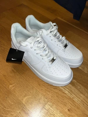 Nike Air Force 1 helvita sneakers oanvända  - Nike Air Force 1 sneakers i helvitt skinn med klassisk siluett, perforerad tå och vit sula. Skorna har snörning, metallbricka med 'AF1' och diskret Nike-logga på sidan. Perfekta för dig som gillar clean och tidlös streetstil.