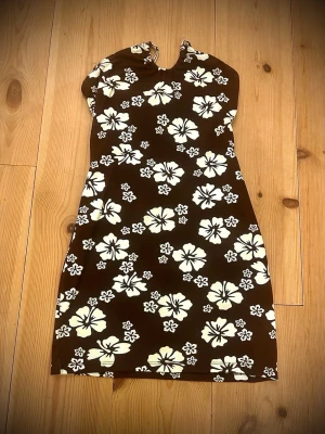 Brun blommig halterneckklänning H&M - Säljer en brun halterneckklänning från H&M Divided i storlek S. Klänningen har ett vitt blommigt mönster och knytning i nacken. Den är kort och har en figurnära passform, perfekt för sommarens varma dagar.