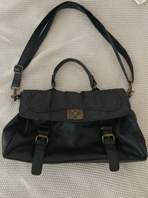 Svart axelväska i skinnlook - Snygg svart axelväska med både handtag och justerbar axelrem. Väskan har mässingsfärgade metalldetaljer, vridlås framtill och två dekorativa spännen. Insidan är fodrad med blommigt tyg och har flera fack med dragkedja. Perfekt för dig som vill ha plats för allt.