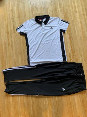 Adidas svartvita träningskläder - Säljer ett set från Adidas med svartvita träningsbyxor och matchande pikétröja. Byxorna har vita ränder längs sidorna och elastisk midja, medan tröjan har svart krage och svarta detaljer längs sidorna. Perfekt för sport eller chill dagar.
