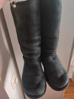 Svarta UGG boots i mocka - Klassiska svarta UGG boots i mjuk mocka med rund tå och platt sula. Insidan är fodrad för extra värme och komfort. Perfekta för kalla dagar och har en enkel, stilren look med UGG-logga bak på hälen.