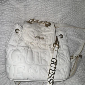 Vit quiltad ryggsäck från Guess - Snygg vit ryggsäck från Guess med quiltad design och stora broderade loggor. Väskan har guldfärgade detaljer, kedja på axelremmen och snörning upptill. Perfekt storlek för det viktigaste och en riktigt trendig accessoar.