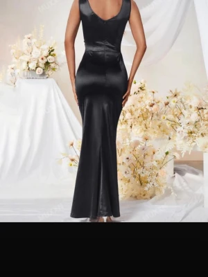 Svart långklänning i satin - Elegant svart långklänning i glansigt satinliknande material. Klänningen har v-ringning, omlottdetalj framtill och en hög slits på sidan som ger en snygg touch. Figurnära passform och är ärmlös, perfekt för en extra snygg look.