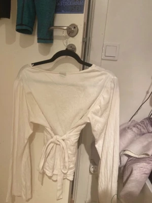 Vit omlottopp med knytband från Gina tricot  - Säljer en vit omlottopp med långa ärmar och knytband i midjan. Toppen har v-ringning och är tillverkad i ett mjukt bomullstyg. Perfekt för dig som gillar en enkel men snygg stil med lite extra detalj i midjan. Storlek m men passar mig som är xs s Skriv för mer frågor❤️