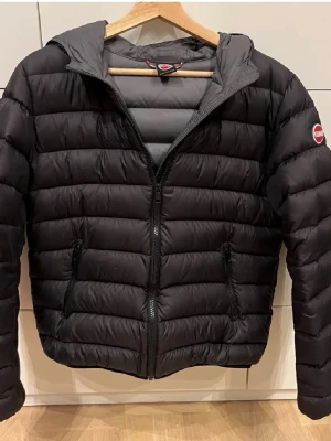 Svart pufferjacka från Colmar, 172cm! - Säljer en svart pufferjacka från Colmar som är storlek 172cm lång eller 16y med huva och dragkedja framtill. Jackan har en quiltad design och två fickor med dragkedja. Perfekt för kyliga dagar och har en stilren look med Colmar-logga på ärmen.