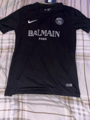 Svart PSG x Balmain Nike t-shirt M - Säljer en svart PSG x Balmain Paris t-shirt från Nike i Dri-FIT material. Tröjan har vita loggor och text framtill, rund hals och korta ärmar. Perfekt för dig som gillar streetwear och sportiga vibes.