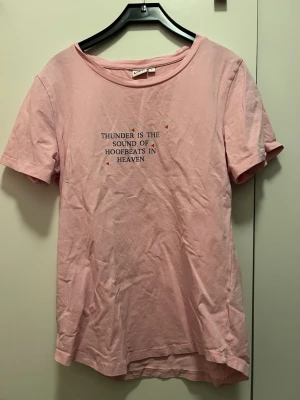 Rosa t-shirt från CRW med tryck - Säljer en ljusrosa t-shirt från CRW i storlek S. T-shirten har rund hals, korta ärmar och ett tryck på bröstet med texten 'Thunder is the sound of hoofbeats in heaven'. Tillverkad i mjuk bomull, perfekt för chill dagar.