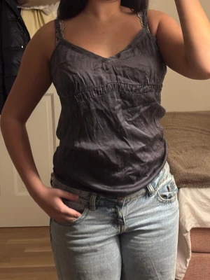 Sidenlinne - Säljer ett mörkgrått linne i siden med tunna axelband och spetsdetaljer. Toppen har v-ringning och en lös passform som ger en avslappnad vibe. Perfekt till jeans eller kjol för en enkel men snygg look.