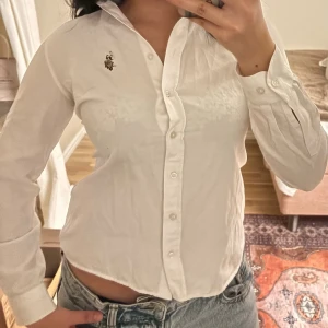 Vit skjorta - Klassisk vit skjorta från med broderad logga på bröstet. Skjortan har lång ärm, knappar framtill och en snygg krage. Perfekt till jeans eller kjol för en clean och stilren look.