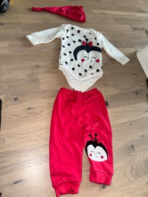 Vit och röd prickig väst med nyckelpiga Nyckelpiga Set - Body, Byxor och Mössa - Säljer en söt vit väst med svarta prickar och ett stort nyckelpigemotiv framtill, dekorerad med en röd rosett. Västen har lång ärm och är gjord i mjuk bomull, perfekt för dig som gillar gulliga detaljer och lekfull stil.Sött set med en vit body med svarta prickar och en söt nyckelpiga applikation. Tillhörande röda byxor med en liknande nyckelpiga applikation på benet. En röd mössa ingår också.