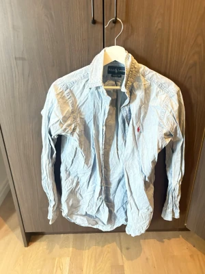 Ljusblå skjorta från Ralph Lauren - Klassisk ljusblå skjorta från Ralph Lauren med röd broderad logga på bröstet. Skjortan har lång ärm, knappar framtill och en tidlös krage. Perfekt till jeans eller chinos för en clean och stilren look.