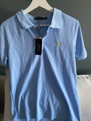 Ljusblå pikétröja från Polo Ralph Lauren - Säljer en klassisk ljusblå pikétröja från Polo Ralph Lauren med gul broderad logga på bröstet. Tröjan har krage, två knappar framtill och är kortärmad. Tillverkad i bomull för en skön och luftig känsla. Perfekt till jeans eller shorts. Säljer pga sent återköp så säljer den för billigare en ny pris. Där vi ser pris och vart jag köpte den ifrån på sista sidan🙂 pris kan diskuteras 