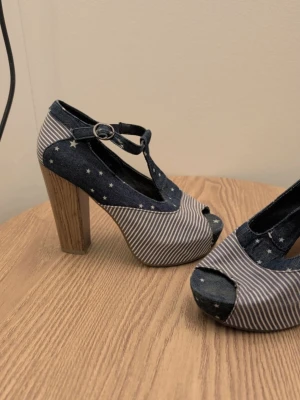 Blå pumps med stjärnor och ränder - Unika pumps i blått denimliknande tyg med vita stjärnor och ränder. Skorna har kraftig träfärgad klack, öppen tå och T-rem med spänne. Plattform framtill för extra höjd och cool retrostil. Perfekta för dig som vill sticka ut!