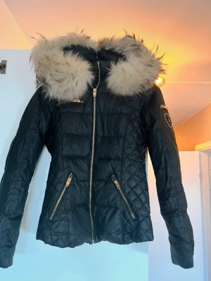 Svart pufferjacka från Rockandblue - Svart pufferjacka med stor fluffig beige fuskpäls på huvan och guldfärgade dragkedjor. Jackan har quiltad design, flera fickor med dragkedja och Rockandblue-märke på ärmen. Perfekt för kalla dagar och har en snygg, modern look.
