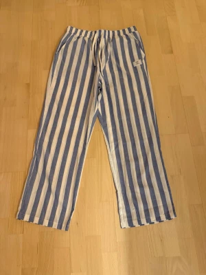 Randiga linnebyxor blå/vit  - Passar L. Säljer ett par chill randiga linnebyxor i blått och vitt från Classic Wear Best Style1979. Byxorna har resårmidja med snörning och raka ben. Perfekta för sommarens lata dagar eller när du vill ha det extra bekvämt.