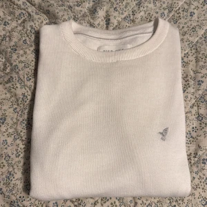  stickad Pier One tröja - Vit stickad tröja från Pier One med rund hals och diskret broderad fågel på bröstet. Tröjan har en klassisk och clean look med ribbade muddar vid halsen. Perfekt för dig som gillar stilrena och enkla plagg.