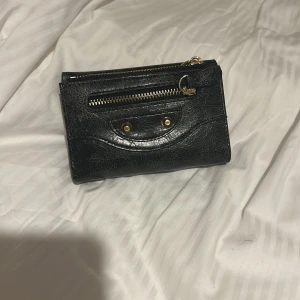 Svart plånbok med gulddetaljer - Snygg svart plånbok i skinn med guldfärgade detaljer och dragkedja framtill. Insidan har flera fack för kort och ett fönster för ID. Stilren design med struktur på ytan och praktisk knappstängning. Perfekt för att hålla koll på alla dina kort och cash. 