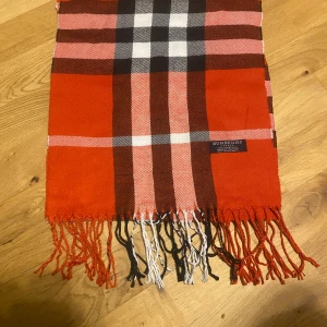  Burberry halsduk - Snygg rutig halsduk från Burberry i 100% lammull. Färgerna är röd, svart, vit och brun med klassiskt rutmönster och fransar i ändarna. Perfekt accessoar för att lyfta din stil under kyliga dagar.