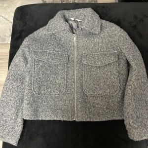 Grå teddyjacka från H&M med dragkedja - Mysig grå teddyjacka från H&M med fluffig textur, två stora bröstfickor och dragkedja framtill. Jackan har en rak, boxig passform och klassisk krage. Perfekt för kyliga dagar när du vill ha en stilren men chill look.