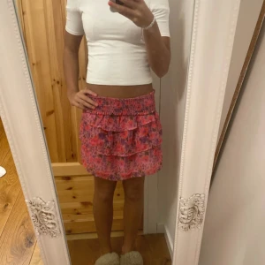 Rosa blommig volangkjol - Supersöt kort kjol i rosa och lila toner med blommigt mönster och bred smock i midjan. Kjolen har flera volanger som ger en fluffig och lekfull look. Perfekt för dig som gillar färg och mönster. Materialet känns lätt och luftigt.