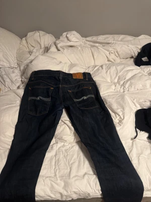 Mörkblå jeans från Nudie Jeans W34 L32 - Säljer ett par mörkblå jeans från Nudie Jeans med klassisk orange kontrastsöm och snygga broderade bakfickor. Modellen har rak passform och normal midja. Materialet är slitstarkt denim i bomull. Perfekt för dig som gillar stilrena och tidlösa jeans.