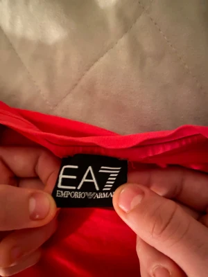 Röd EA7 Emporio Armani t-shirt - Snygg röd t-shirt från EA7 Emporio Armani med vit logga på bröstet och svart/vit logoband längs sidan. Klassisk passform och rund hals. Tillverkad i mjuk bomull som känns skön mot huden. Perfekt för dig som gillar streetwear och vill sticka ut.