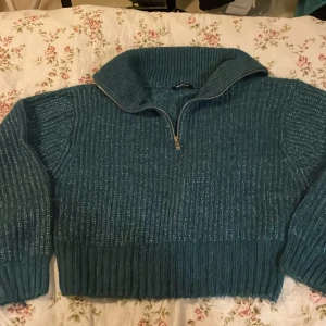 Blågrön stickad half zip tröja Gina Tricot - Mysig blågrön  stickad tröja från Gina Tricot i storlek S. Tröjan har en bred krage och half zip-dragkedja framtill. Ribbad struktur och långa ärmar ger en chill och trendig look. Perfekt för kyliga dagar när du vill vara både bekväm och snygg.Midje modell.