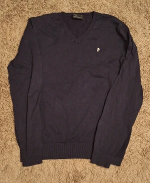 Mörkblå v-ringad tröja Peak Performance - Snygg mörkblå v-ringad tröja från Peak performance med diskret logga på bröstet. Tröjan är i bomull och har långa ärmar och ribbade muddar vid ärmslut och nederkant. Perfekt för dig som gillar stilrena och klassiska plagg.