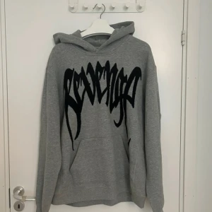 Grå Revenge hoodie med svart tryck - Hoodie från Revenge. Haft den undanlagd så är i super bra skick, tyvärr har man växt ut storleken den så säljer vidare.