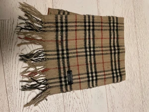Rutig beige halsduk från Burberry - Klassisk halsduk från Burberry i beige med rutigt mönster i svart, vitt och rött. Gjord av 100% kashmir för en lyxig och mjuk känsla. Halsduken har fransar i ändarna och den ikoniska Burberry-taggen. Perfekt accessoar för att lyfta din stil.
