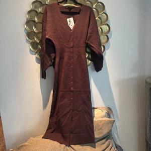 Brun långklänning med knappar constrictors - Snygg brun långklänning från Selected Femme med v-ringning och knappar hela vägen framtill. Klänningen har trekvartsärmar och är gjord i mjukt stickat material som känns skönt mot huden. Perfekt för dig som gillar stilrena och enkla plagg.