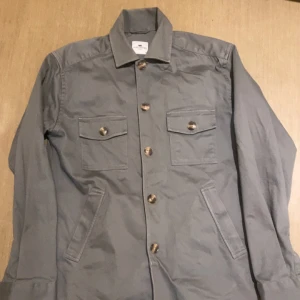 Grå/grön overshirt från John Henric XS - Snygg grå/grön overshirt från John Henric i storlek XS. Jackan har klassisk krage, två bröstfickor med lock och knappar samt snedställda sidofickor. Stängs med stora knappar framtill och är perfekt för lager-på-lager-stil.