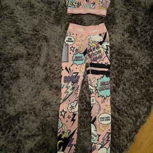 Stronger leggings med pop art-mönster - Säljer ett par färgglada leggings från Stronger med pop art-inspirerat mönster i rosa, blått, svart och gult. Hög midja och tight passform, perfekta för träning eller chill. Snygga detaljer med text och grafiska element som gör dem unika.