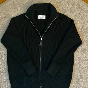 Svart ribbad kofta från Maison Margiela - Svart ribbstickad kofta från Maison Margiela med hög krage och hel dragkedja framtill. Ikoniska vita stygn i nacken och grovstickad känsla. Perfekt för dig som gillar minimalistisk och clean stil. Snygg att bära över en t-shirt eller skjorta.