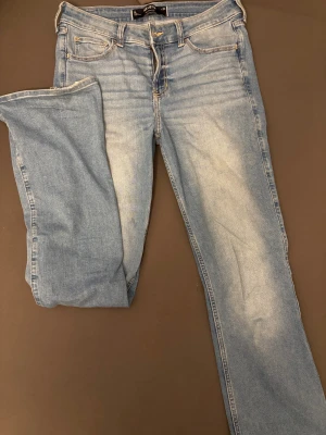 Ljusblå bootcut jeans från Hollister - Säljer ett par ljusblå midwaist bootcut jeans från Hollister med klassisk femficksdesign och snygga kontrastsömmar på bakfickorna. Jeansen har normal passform och är tillverkade i mjukt denim med lätt tvättad look. Perfekt för dig som gillar avslappnad stil.