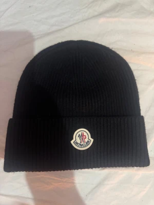 Moncler mössa - Svart ribbstickad mössa från Moncler med klassisk uppvikt kant och snyggt Moncler-märke framtill. Perfekt för dig som vill ha en clean och stilren look under kalla dagar. Materialet är mjukt och värmande, och mössan har en enkel, tidlös design. Var inget för mig, testad 1 gång inomhus