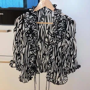 Zebramönstrad bolerotopp med volanger - Säljer en svartvit bolerotopp med zebramönster och volangkant längs halsringning, ärmslut och nederkant. Tunn och luftig, perfekt för att ge outfiten extra edge. Knytband framtill och trekvartsärmar. Riktigt snygg att styla över linne eller klänning.