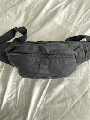 Svart magväska från Urban Classics - Snygg svart magväska från Urban Classics med justerbart band och praktisk dragkedja framtill. Perfekt för att ha med sig det viktigaste och hålla händerna fria. Väskan är tillverkad i slitstark polyester och har en stilren, enkel design.