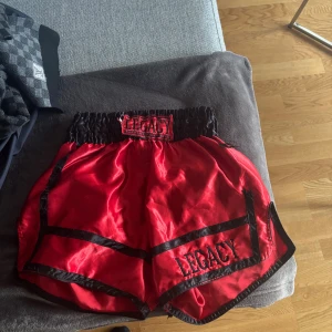 Röda Legacy boxningsshorts - Snygga röda muaythai shorts från Legacy med svarta detaljer och broderad logga. De har bred resår i midjan och är tillverkade i glansigt material som ger en sportig vibe. Perfekta för träning eller match.  (thaiboxning) liten skada vid texten på midjan  