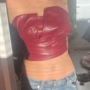 Vinröd tubtopp i skinnimitation - Säljer en vinröd korsett tubtopp i skinnimitation med snygga veckade detaljer framtill. Toppen har en mjuk insida och en dragkedja baktill för enkel på- och avtagning. 