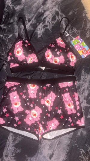 Care Bears pyjamas set med rosa nallemönster - Supergullig pyjamas från Care Bears i storlek S med svart botten och rosa nallebjörnar, stjärnor och hjärtan över hela tyget. Toppen har smala axelband och byxan är i shortsmodell. Materialet är mjukt och stretchigt, 90% polyester och 10% elastan. Perfekt för dig som älskar söta prints!