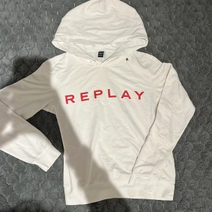 Vit hoodie från Replay med röd glittertext - Vit hoodie från Replay med stor röd glittrig text på bröstet. Tröjan har huva, långa ärmar och ribbade muddar. Materialet är mjuk bomull och insidan är lätt borstad för extra komfort. 