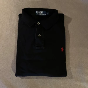 Ralph Lauren pikétröja långärmad  - Svart pikétröja från Polo Ralph Lauren med klassisk krage och två vita knappar framtill. Tröjan har det ikoniska röda Polo-logot broderat på bröstet. Tillverkad i mjuk bomull som är skön mot huden och passar perfekt till jeans eller shorts.