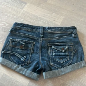 Egna gjorda Blå jeansshorts med detaljerade fickor - Säljer ett par blå jeansshorts med coola detaljerade fickor och snygga nitar. Shortsen har uppvikta benslut, bälteshällor och knappar i metall. Passformen är normal . Perfekt för dig som gillar streetstyle och vill ha något som sticker ut.