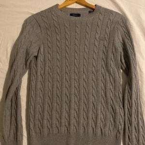 Grå kabelstickad tröja från GANT - Snygg grå kabelstickad tröja från GANT i storlek S. Tröjan har rund hals, ribbade muddar och ett broderat GANT-emblem på bröstet. Perfekt för dig som gillar klassisk stil med en modern touch. Köpt på nordiska kompaniet för 1500kr, mitt pris 700kr