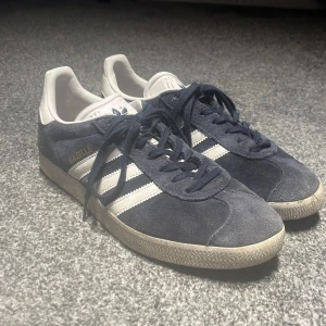 Adidas Gazelle mörkblå sneakers - Säljer ett par klassiska Adidas Gazelle sneakers i mörkblå mocka med vita detaljer och snörning. Skorna har de ikoniska tre ränderna på sidan och en platt sula med mönster. Nypris 900-950 kr. Inga defekter över huvud taget. 